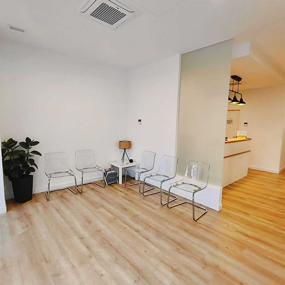 Sala de espera moderna y luminosa de clínica sanitaria con sillas transparentes, suelo de madera y acceso a recepción.